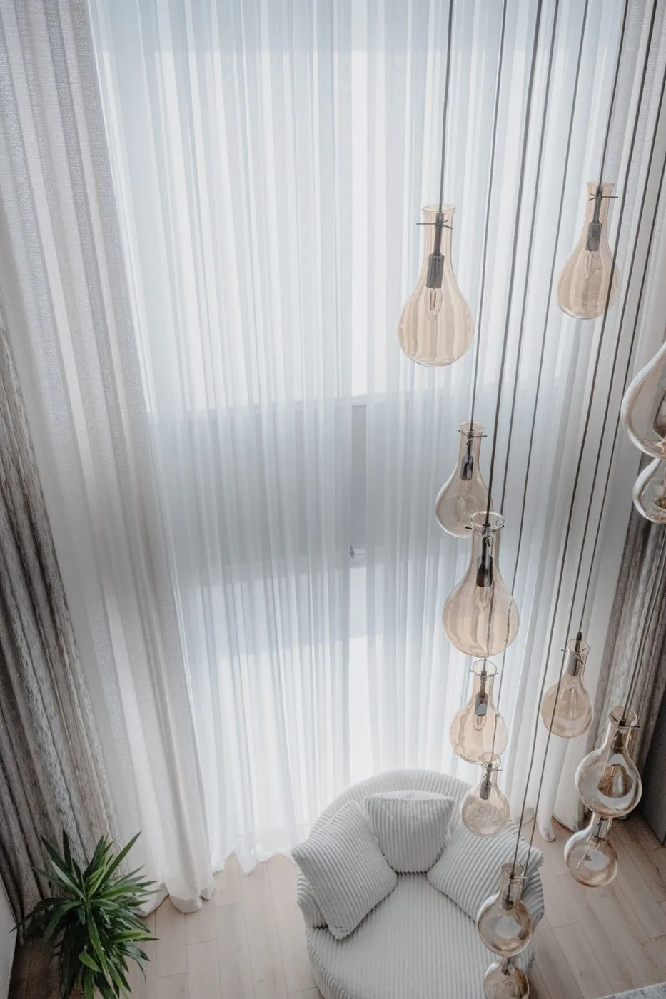 A pale upholstered armchair beside floor-to-ceiling sheer curtains, below a cluster of amber glass pendant lights